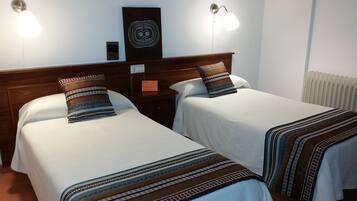 Habitación de hotel con dos camas individuales, cada una con una colcha estampada y una almohada decorativa. Hay luces fijadas en la pared sobre cada cama y una mesita entre ellas.