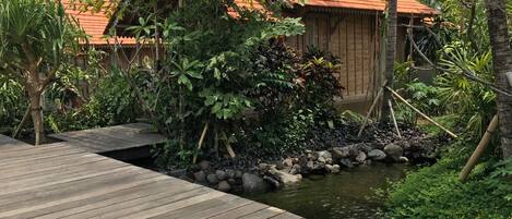 Une terrasse en bois mène à un petit étang bordé de rochers et entouré d’une végétation luxuriante.