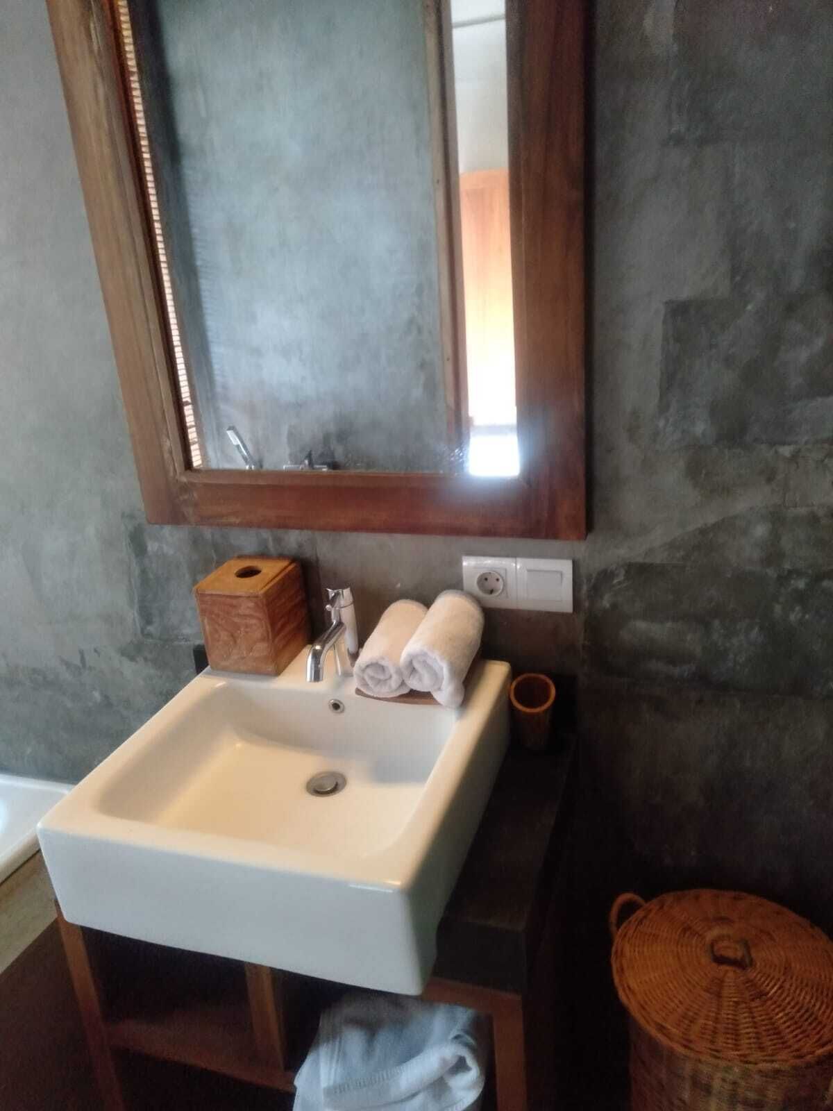 Casa de banho com lavatório branco, um banquinho de madeira, cesto de fibra natural e espelho com moldura de madeira.