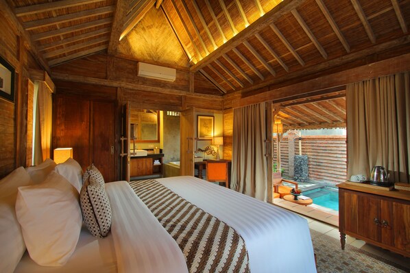 A spacious bedroom with a large bed, wooden ceiling beams, a bathroom with a sink and mirror, and a view of an outdoor pool area.