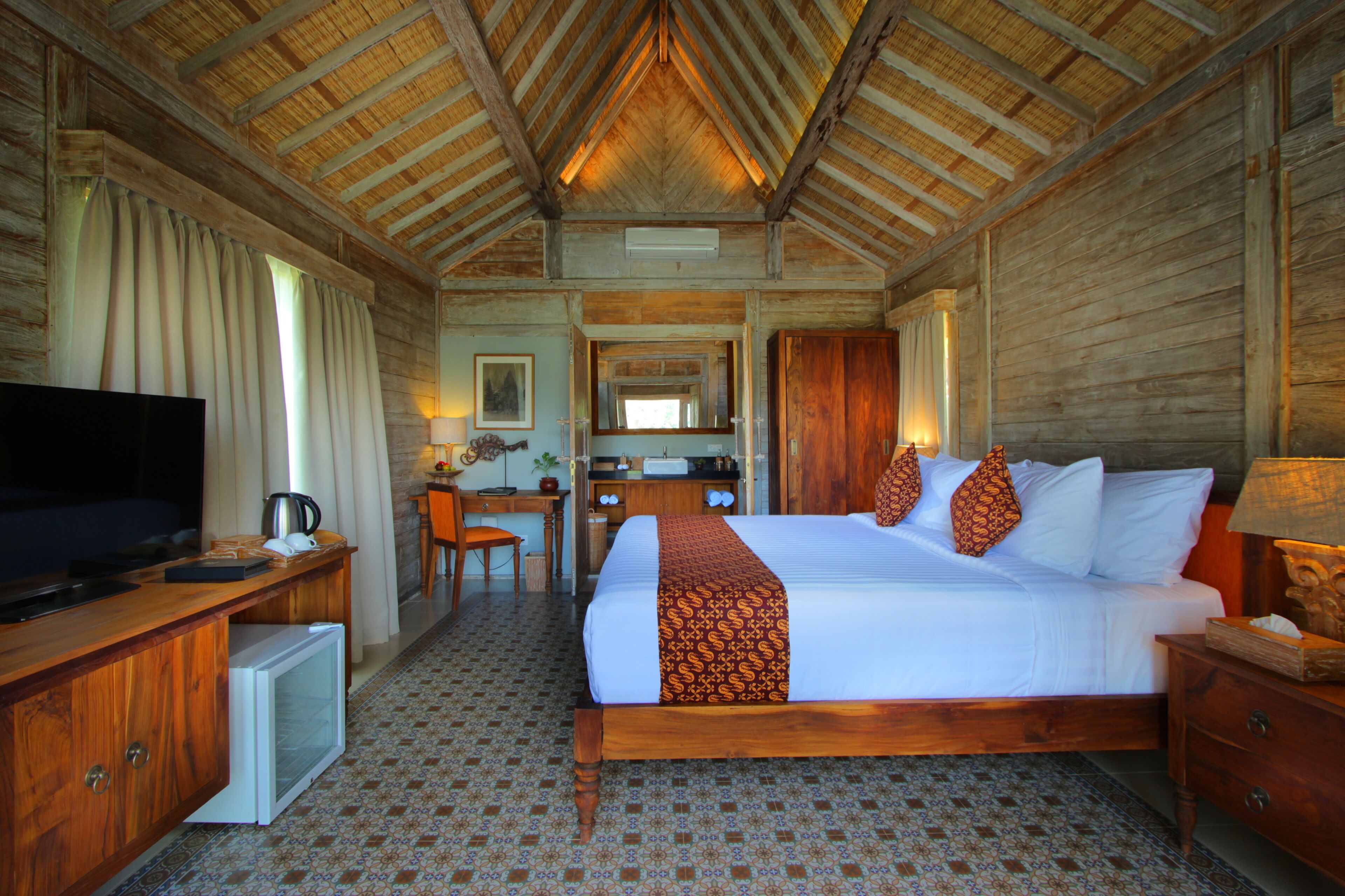 Una habitación de hotel con techo de madera, una cama con ropa de cama estampada, un escritorio con televisor y un refrigerador pequeño.