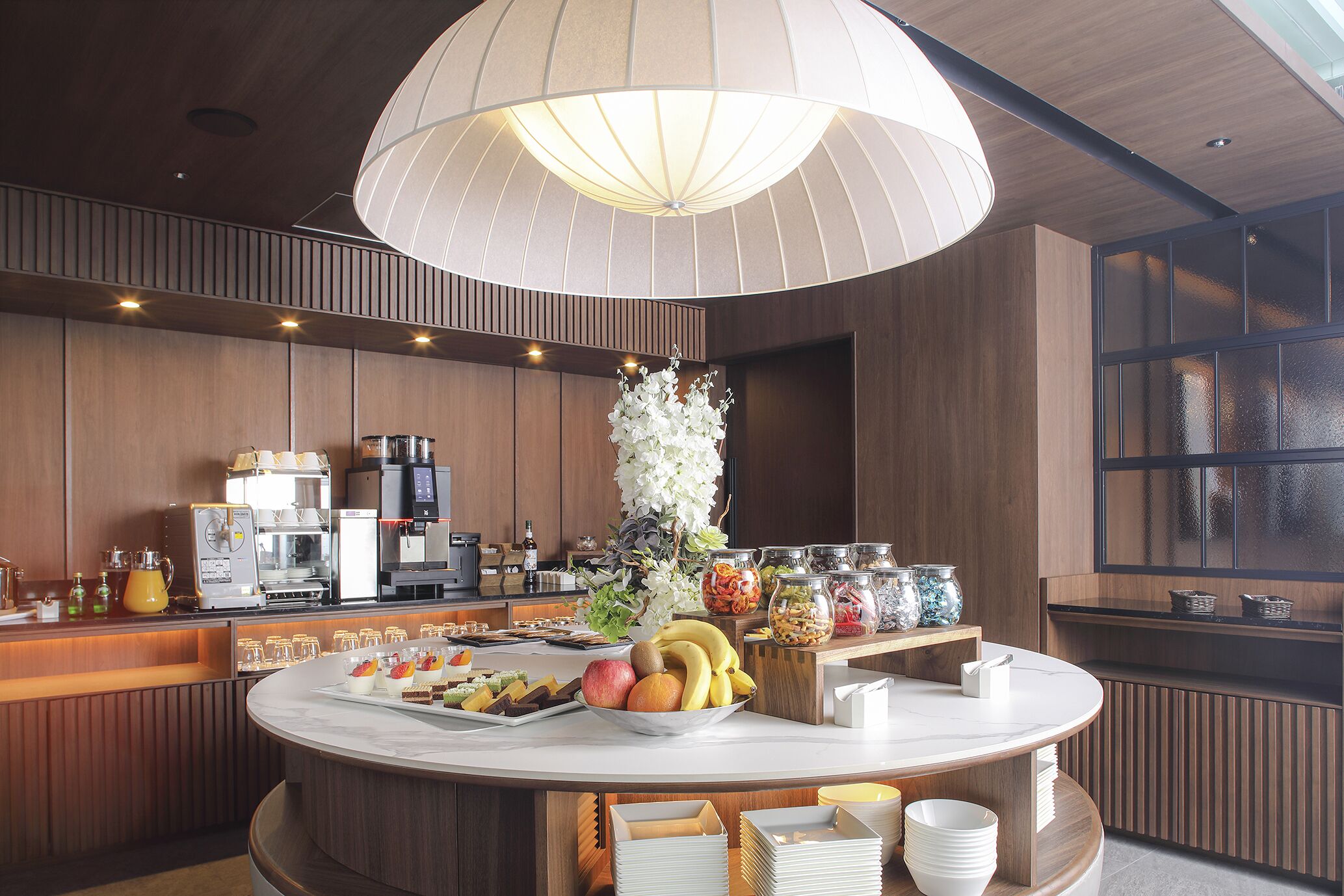 A modern kitchen with a central island, coffee machines, and a variety of food items.