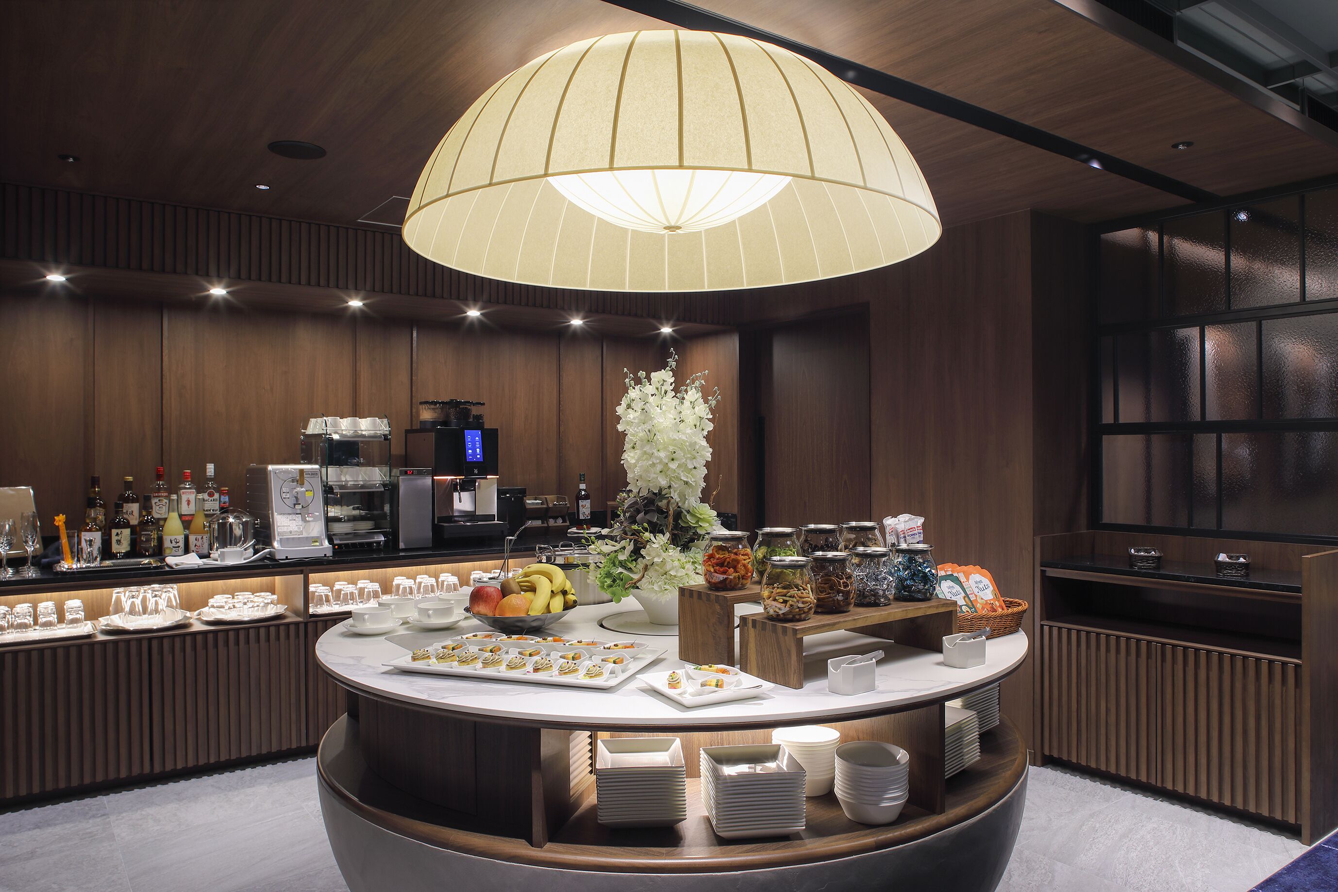 A well-lit buffet area with a central serving station, various food items, and a coffee machine.