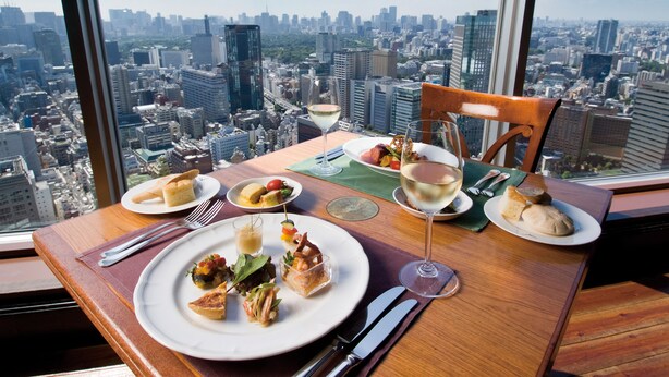 A dining table with plates of food and wine glasses, set against a cityscape view from a high-rise building. - Tokyo Dome Hotel (Tokyo)