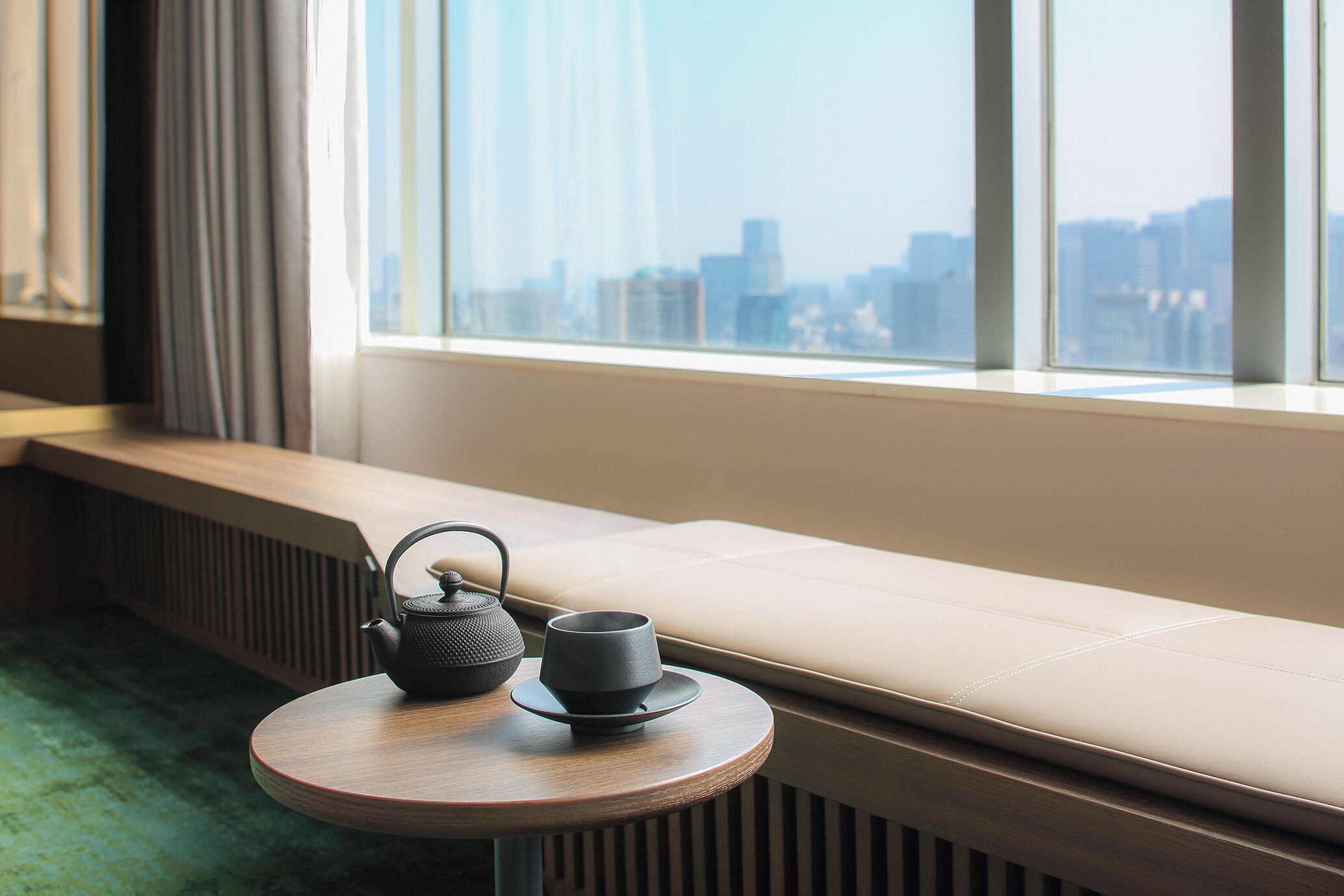 A room with a window seat, a round wooden table, a black teapot, and a cup.