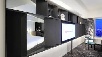 A modern hotel room with a large bed, a TV screen displaying the hotel's name, a seating area with a chair, and a cityscape view.