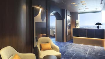 Um hall de hotel moderno com teto alto, carpete azul e área de estar com duas poltronas e uma mesa pequena.