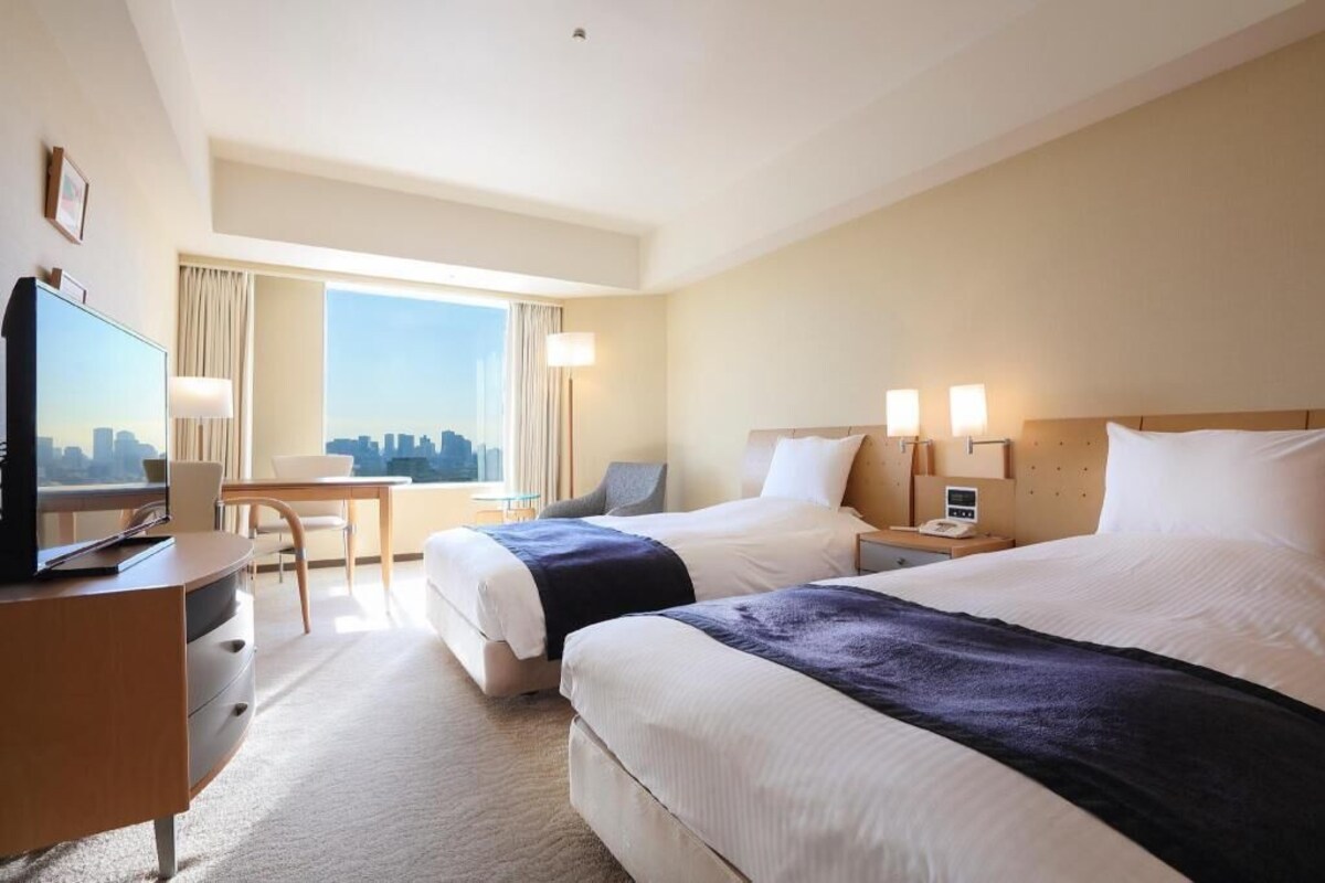 A hotel room with two beds, a desk with a chair, a television, and a city view.