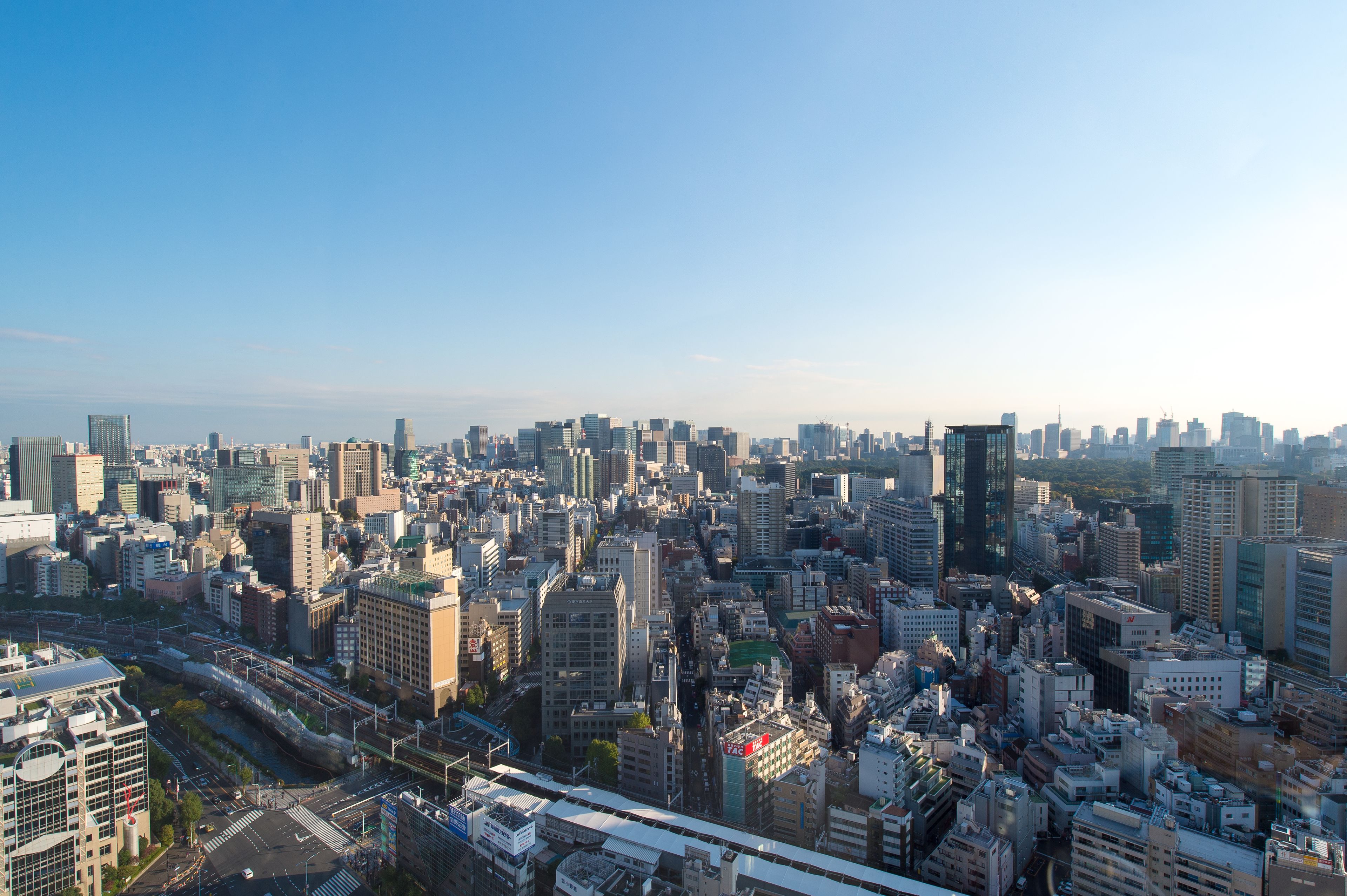 A cityscape with a dense cluster of high-rise buildings, a prominent train track, and a clear sky.