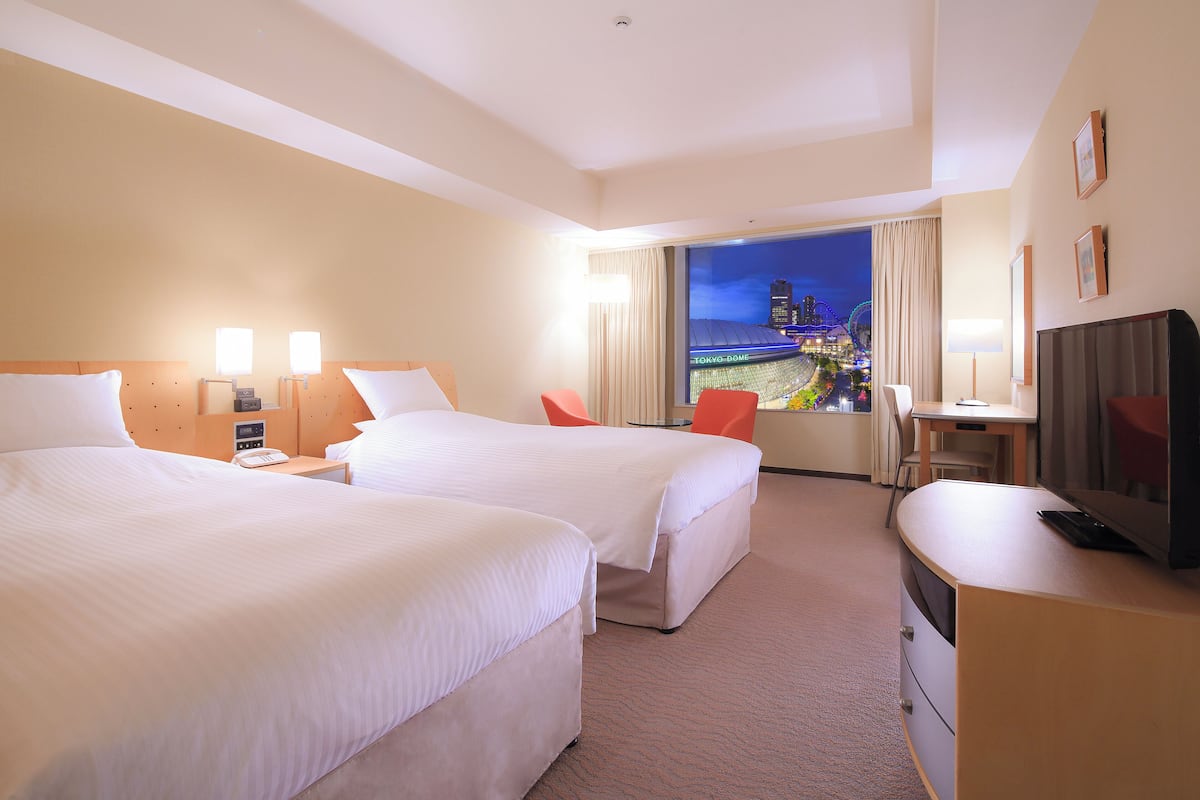 A hotel room with two beds, a desk, a chair, a TV, and a view of Tokyo Dome.