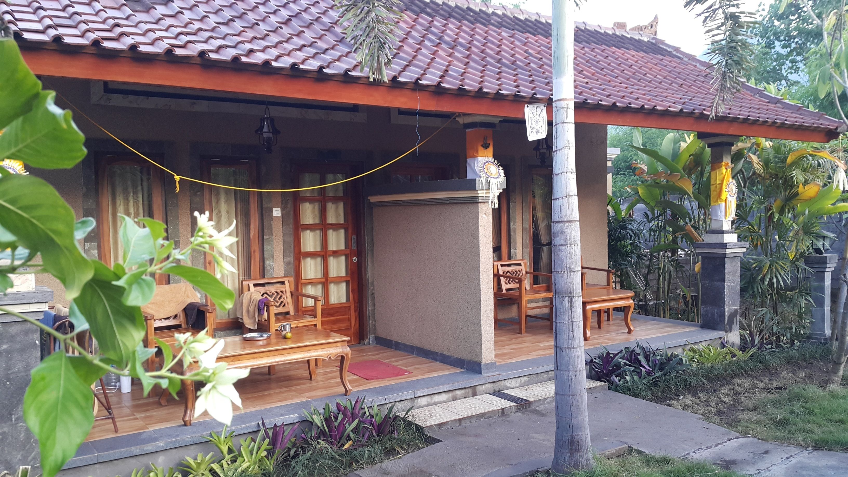 A traditional wooden house with a tiled roof, a porch with wooden furniture, and a small garden area with tropical plants.