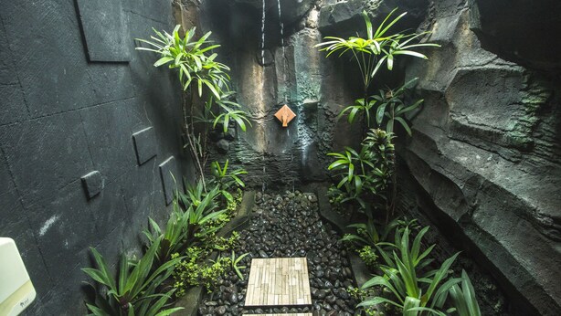 A narrow, rocky indoor garden with a small waterfall, surrounded by lush green plants and a wooden bench. - Trijaya Guest House (Pemuteran)