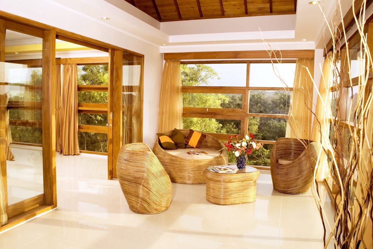 A spacious living area with a large window, a sofa, two woven chairs, and a small round table with a vase of flowers.