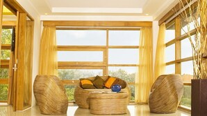 Um quarto espaçoso com teto de madeira alto, grandes janelas e área de estar central com um sofá e duas poltronas.