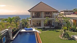 Uma casa de dois andares com piscina, rodeada por vegetação exuberante e vista para o oceano.