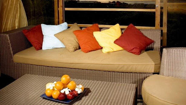 Quarto com uma janela ampla com vista para o pôr do sol, um sofá com almofadas e uma mesa pequena com frutas.