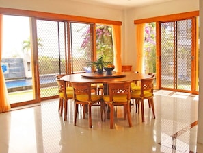 A dining area with a round table and chairs, large windows with curtains, and a view of a pool and garden.