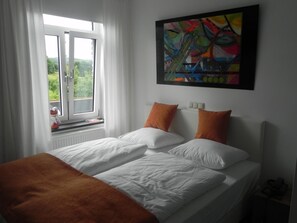 Un dormitorio con cama, ropa de cama blanca, dos almohadas naranjas, una ventana con cortinas blancas y un cuadro abstracto de colores en la pared.