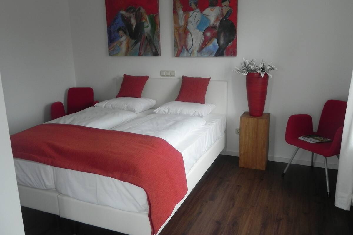 A bedroom with a double bed, two red chairs, a wooden nightstand, and two paintings on the wall.