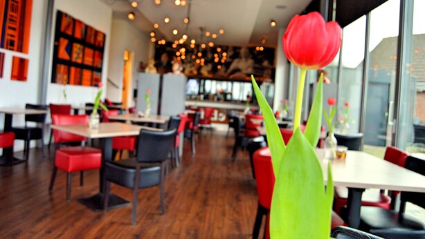 A restaurant interior with red and black chairs, wooden tables, and a large window with a red tulip in the foreground. - Hotel Restaurant Uit de Kunst (Vijlen)