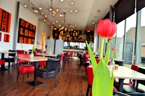 A restaurant interior with red and black chairs, wooden tables, and a large window with a red tulip in the foreground. - Hotel Restaurant Uit de Kunst (Vijlen)