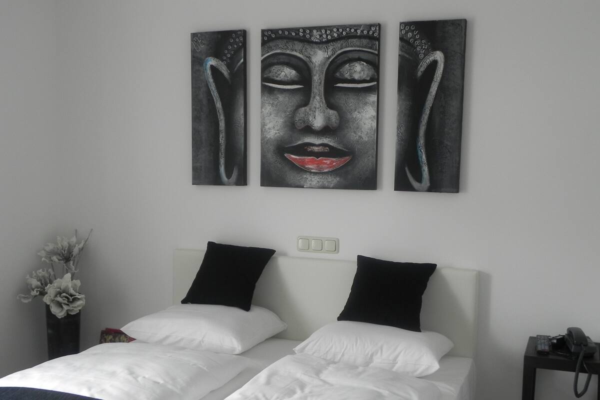 A hotel room with a bed, two black pillows, a white headboard, a nightstand with a phone, and a wall-mounted artwork.