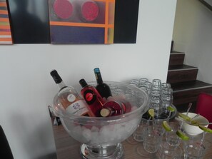 Zona de bar con un recipiente grande lleno de hielo y botellas de alcohol, rodeada de vasos vacíos y una escalera al fondo.