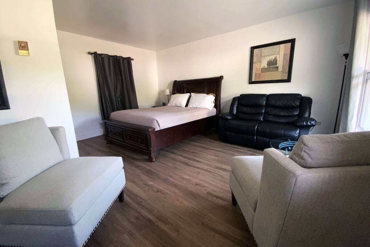 A hotel room with a bed, two armchairs, a black leather sofa, a wooden headboard, a framed picture on the wall, and a window with curtains.