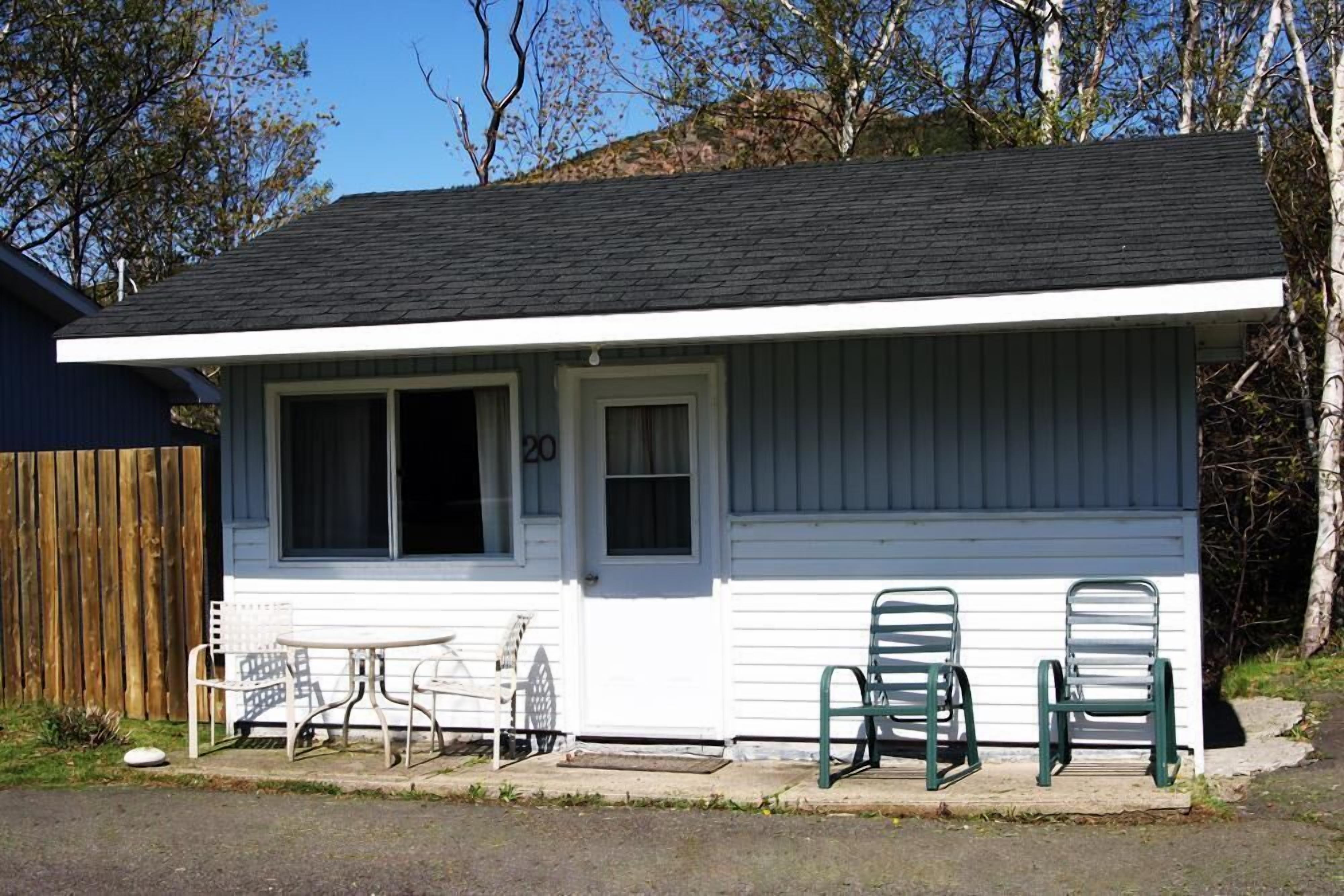 A small, single-story house with a white exterior and a dark roof. There are two windows, one with a curtain and one without. In front of the house, there are two green chairs and a white table.