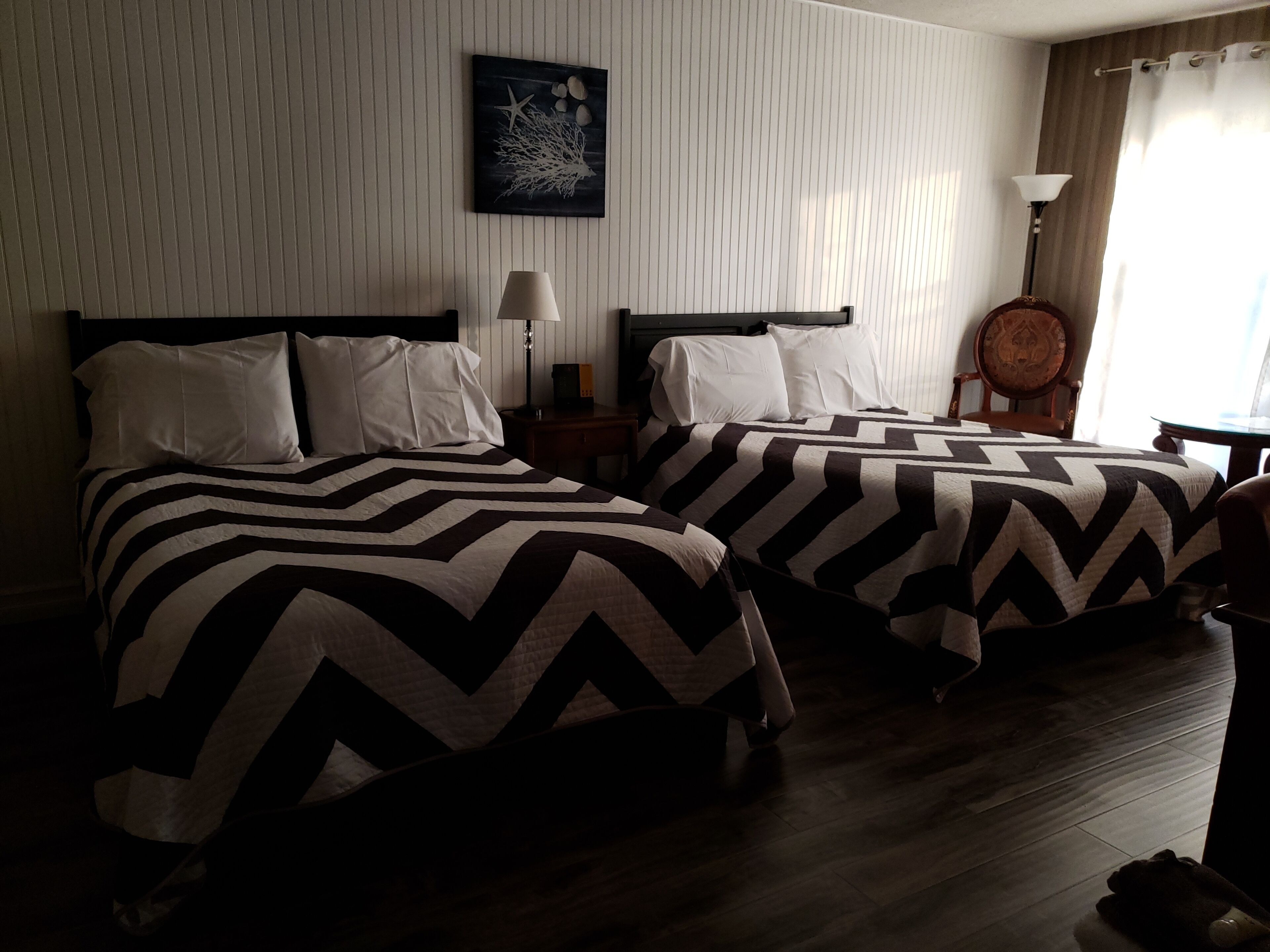 A hotel room with two beds, a nightstand, a lamp, a decorative wall piece, and a wooden chair.