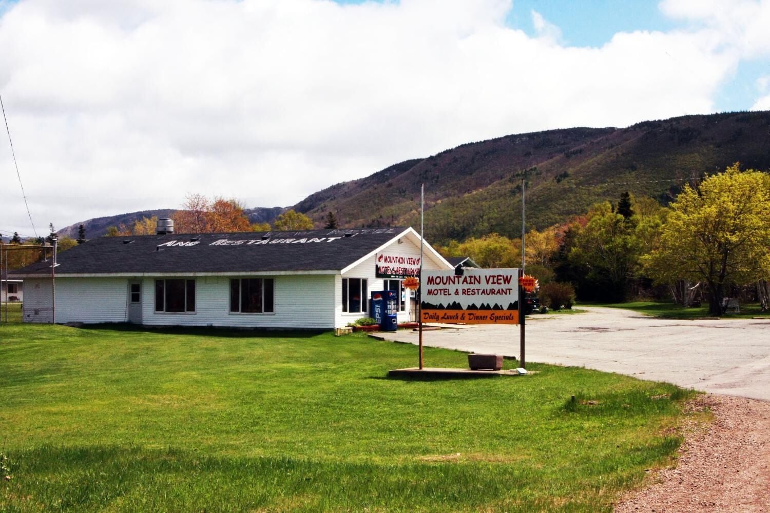 Een motel en restaurant genaamd Mountain View, met een eenvoudig wit gebouw en een zwart dak, een Pepsi-koelkast en een bord waarop dagelijkse lunch- en dinerspecials worden aangeboden.