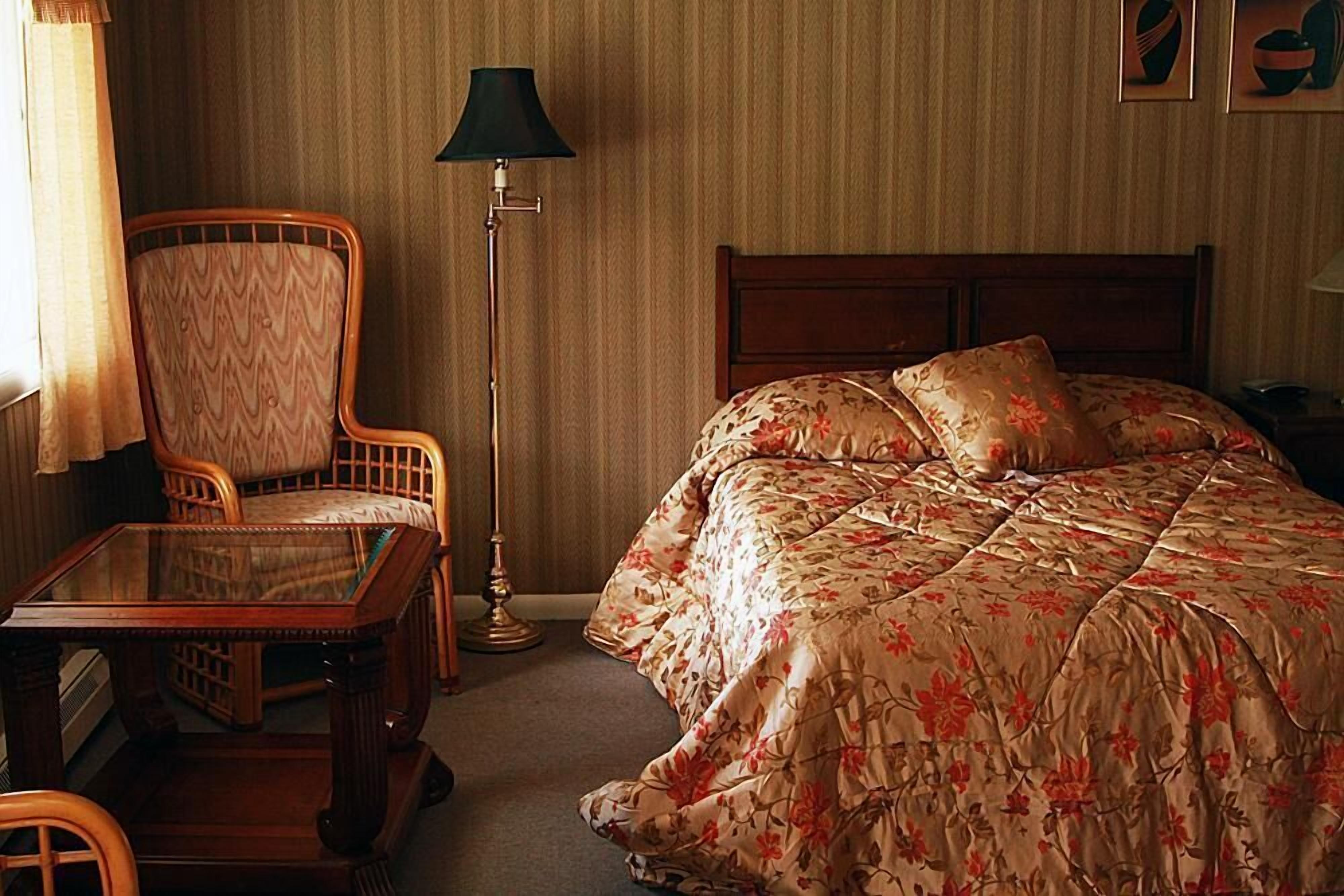 Habitación de hotel con una cama de madera, ropa de cama floral, una silla de madera, una mesa de vidrio y una lámpara de pie.