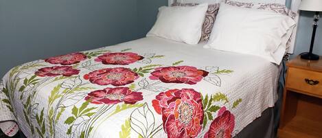 Un lit recouvert d’une couvre-lit à motifs floraux, avec des oreillers blancs et une table de chevet en bois sur laquelle se trouve une lampe.