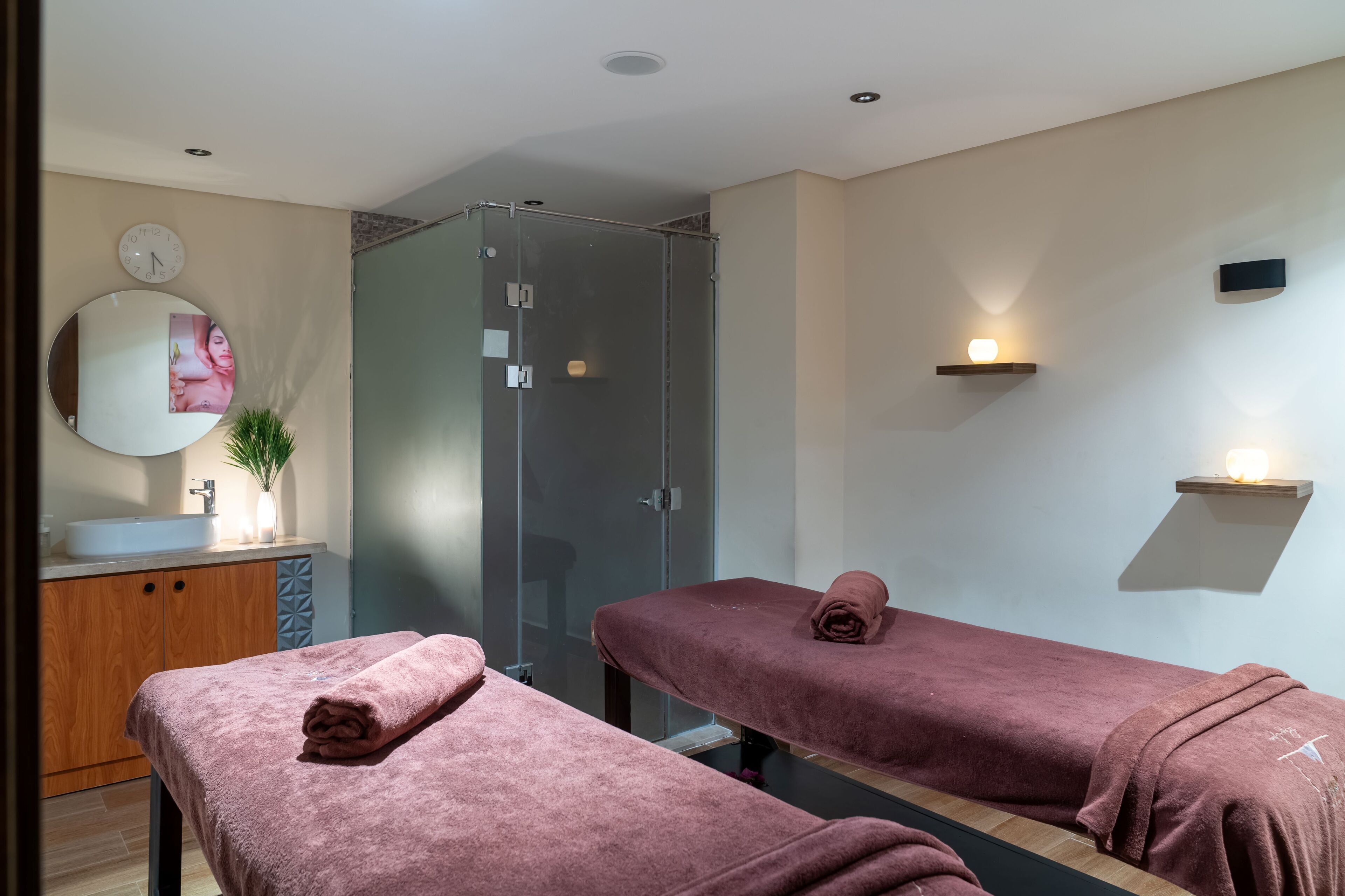 Ein moderner Spa-Raum mit zwei Massagetischen, einer Duschkabine aus Glas, einem Waschbecken und einem Spiegel mit Uhr.