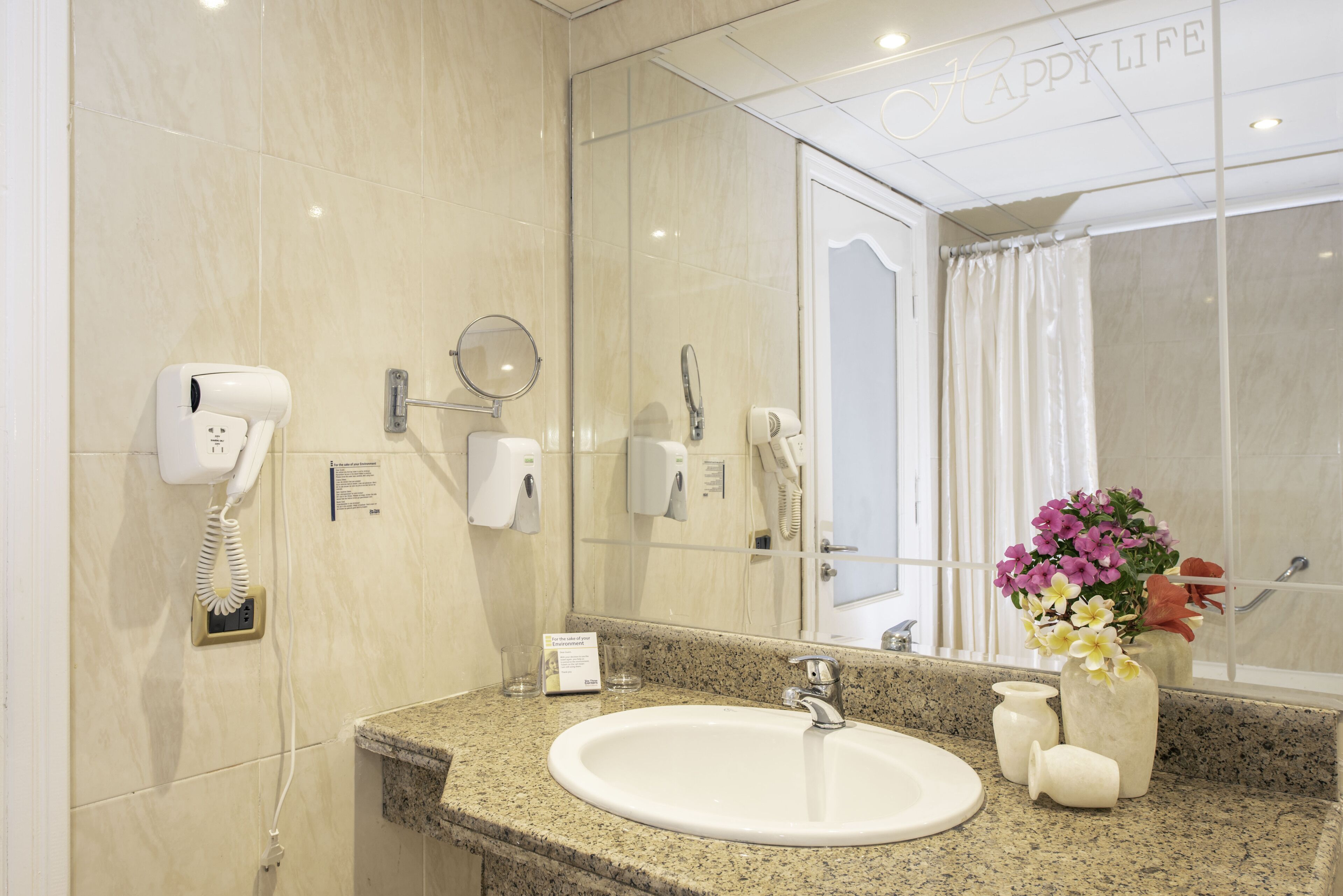 Bagno d'hotel con lavabo su piano in granito, uno specchio, un asciugacapelli, un telefono e un vaso con fiori.