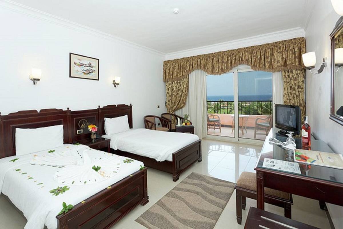 A hotel room with two beds, a desk with a television, a balcony with a view, and a painting on the wall.
