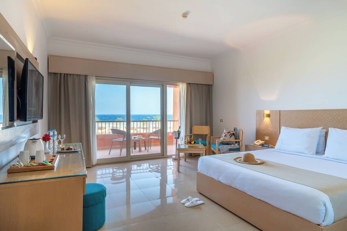 A hotel room with a bed, a desk with a laptop, a chair, a TV, and a balcony with a view of the sea.