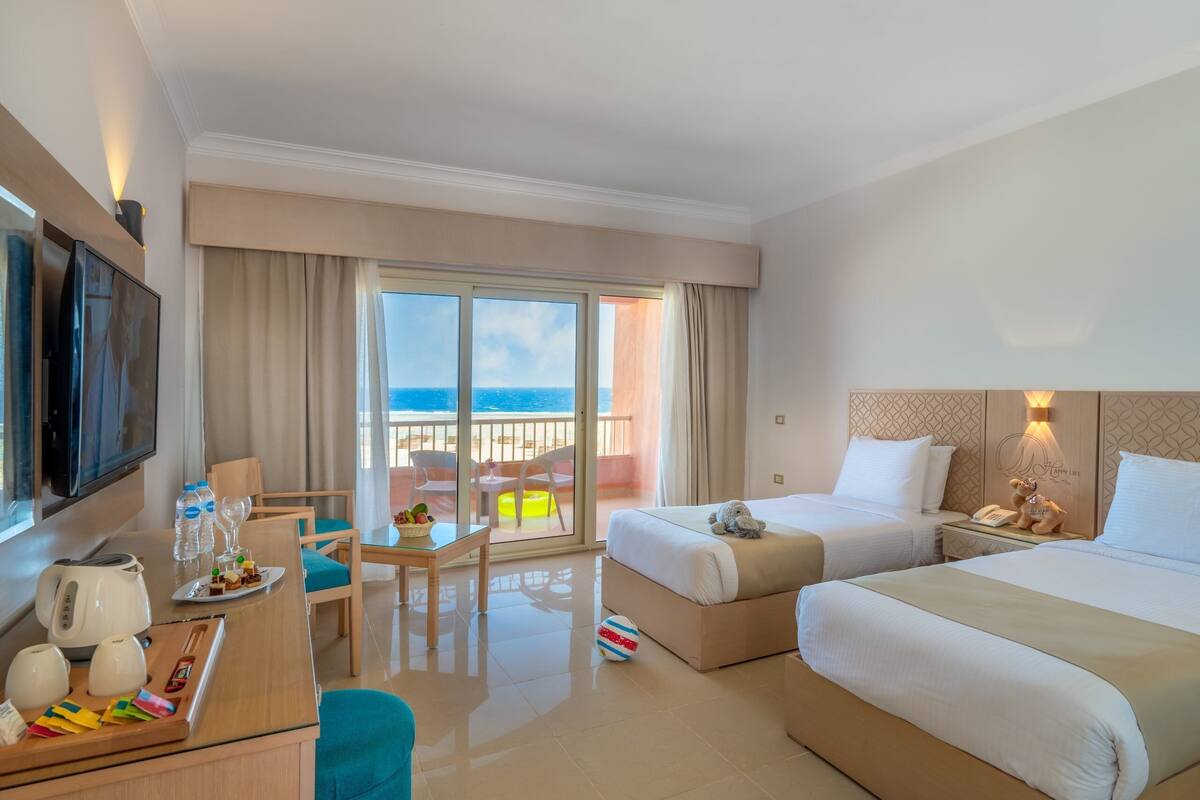 A hotel room with two beds, a TV, a desk with a kettle, a table with a glass of water, and a balcony with a view of the ocean.
