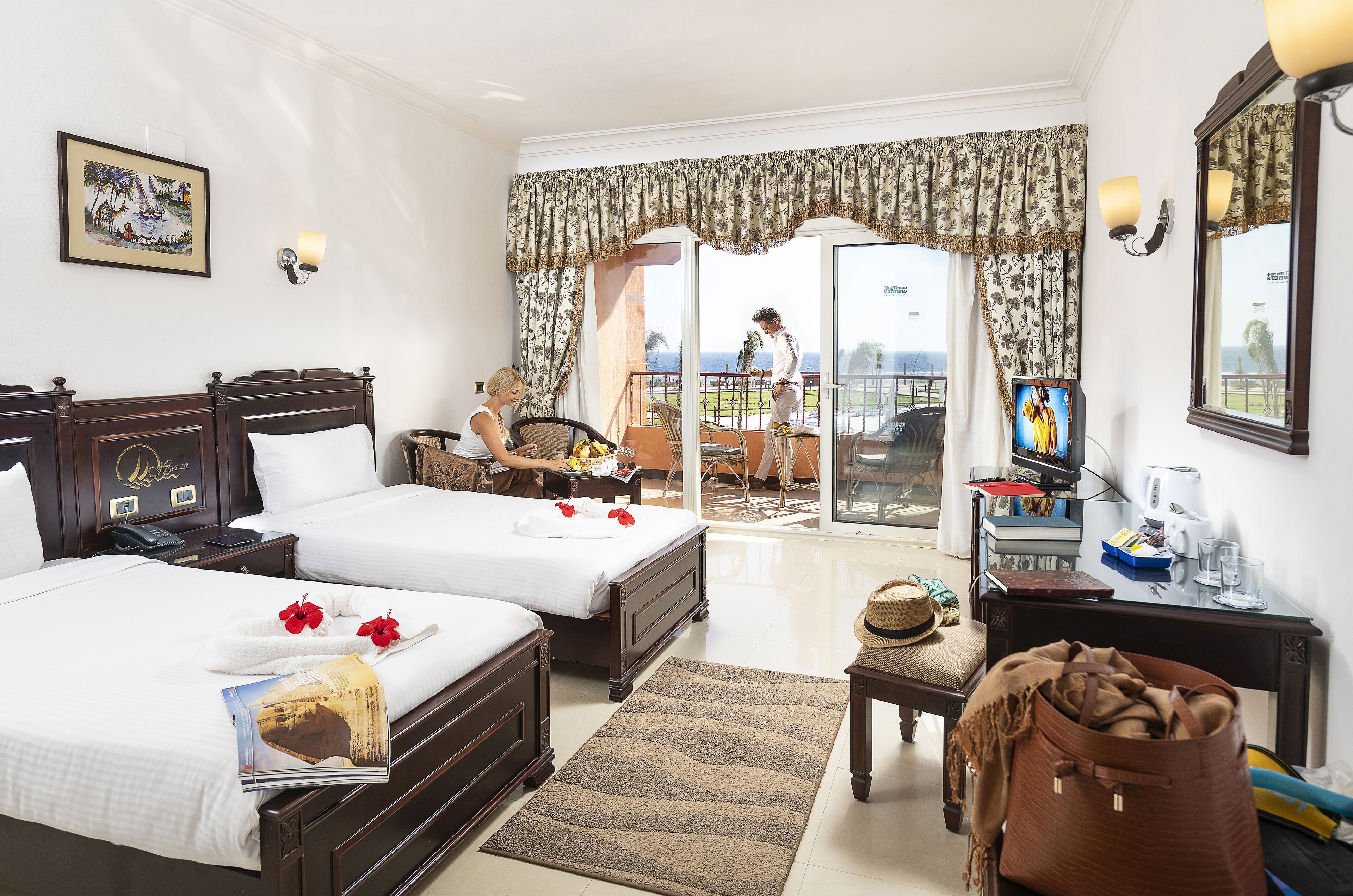 A hotel room with two beds, a balcony, a television, and a dining area.
