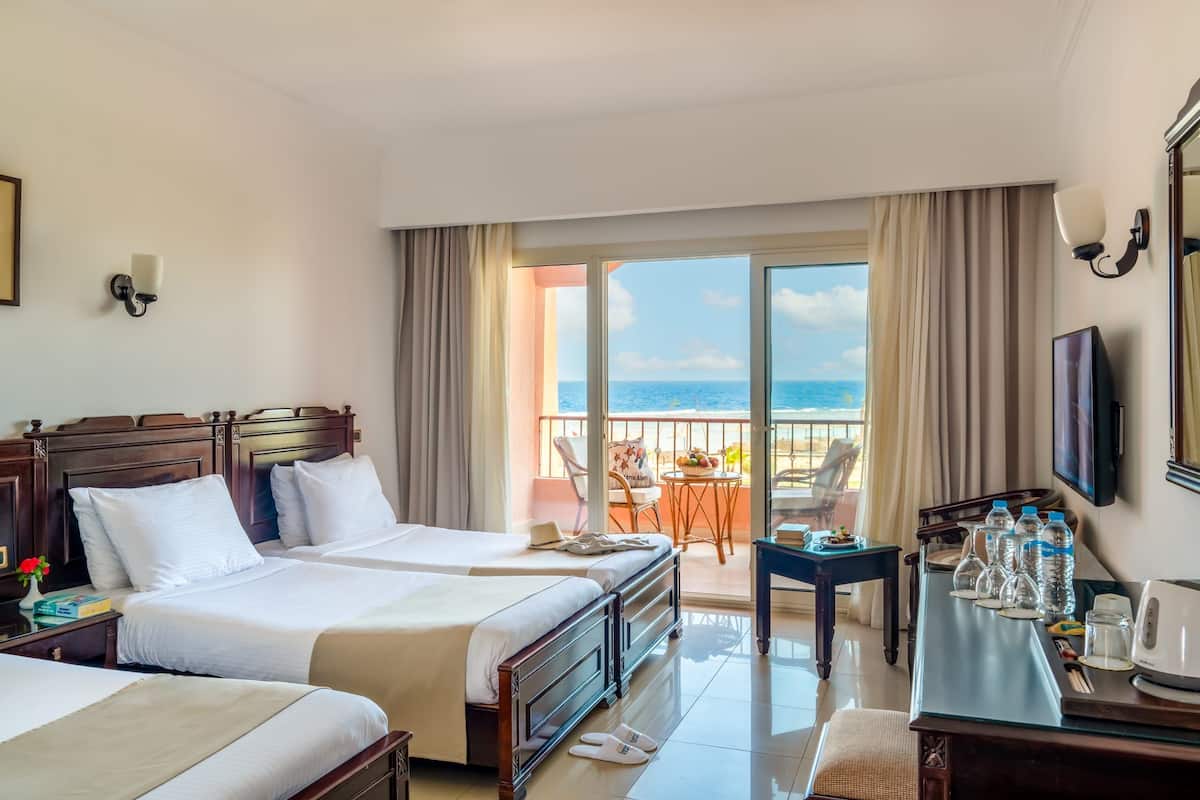 A hotel room with two beds, a balcony with a table and chairs, and a desk with water bottles and a kettle.