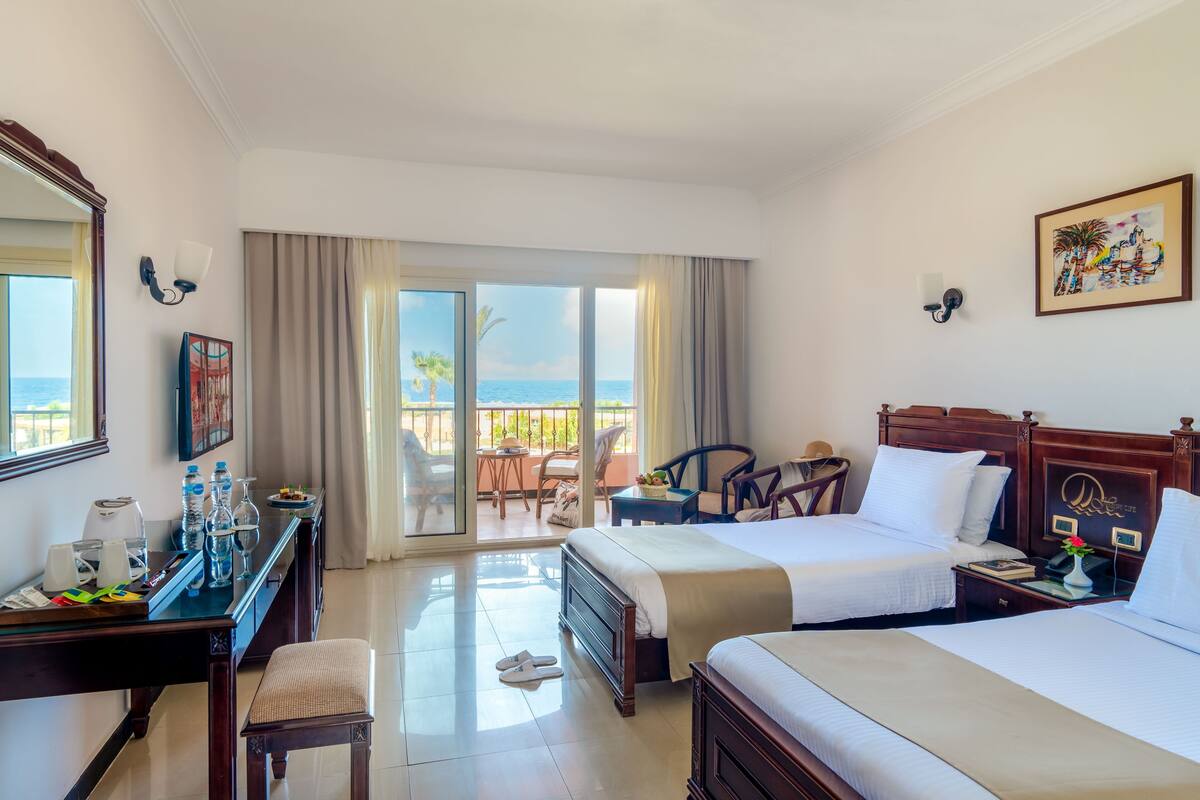 A hotel room with two beds, a desk with a chair, a small table, and a balcony with a view of the ocean.