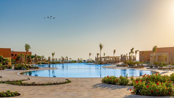 A large outdoor swimming pool with multiple lanes, surrounded by lounge chairs and palm trees, with a clear sky above. - Three Corners Happy Life Beach Resort - All Inclusive (Marsa Alam)