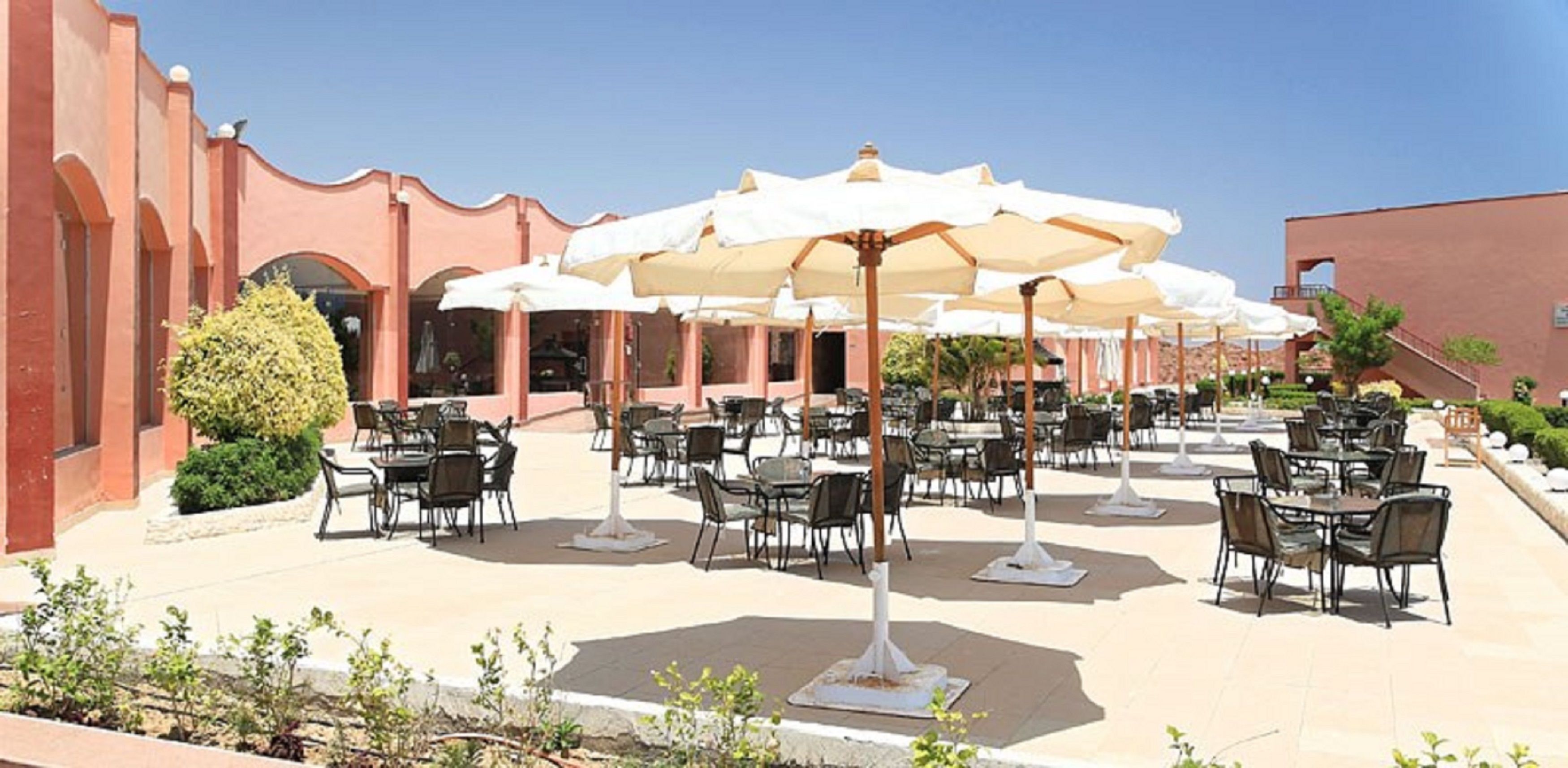 Un área de comedor al aire libre con mesas y sillas, cubierta por sombrillas grandes, y un edificio con un estilo arquitectónico distintivo al fondo.