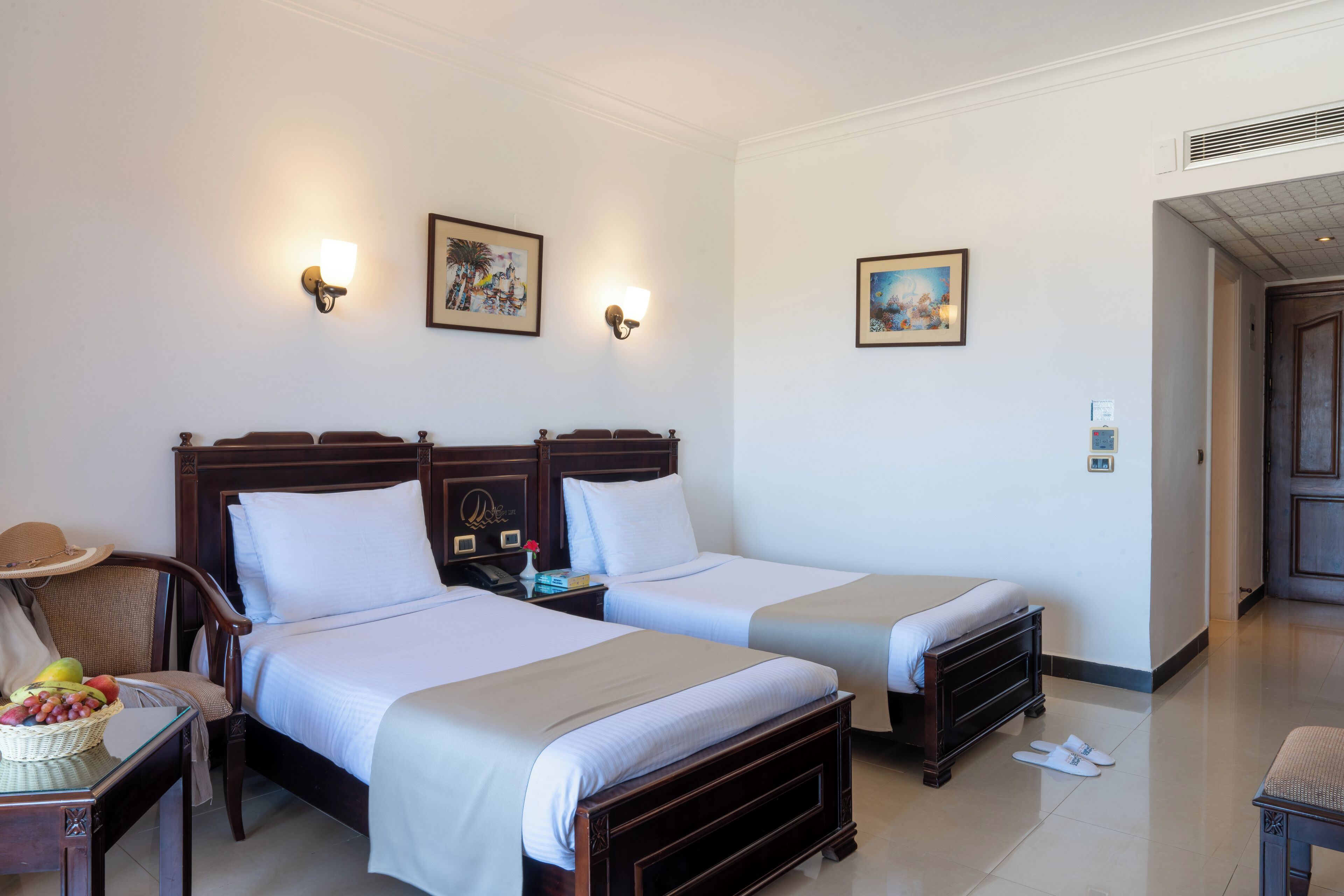 A hotel room with two beds, a nightstand with a lamp, a chair, and a table with fruit.