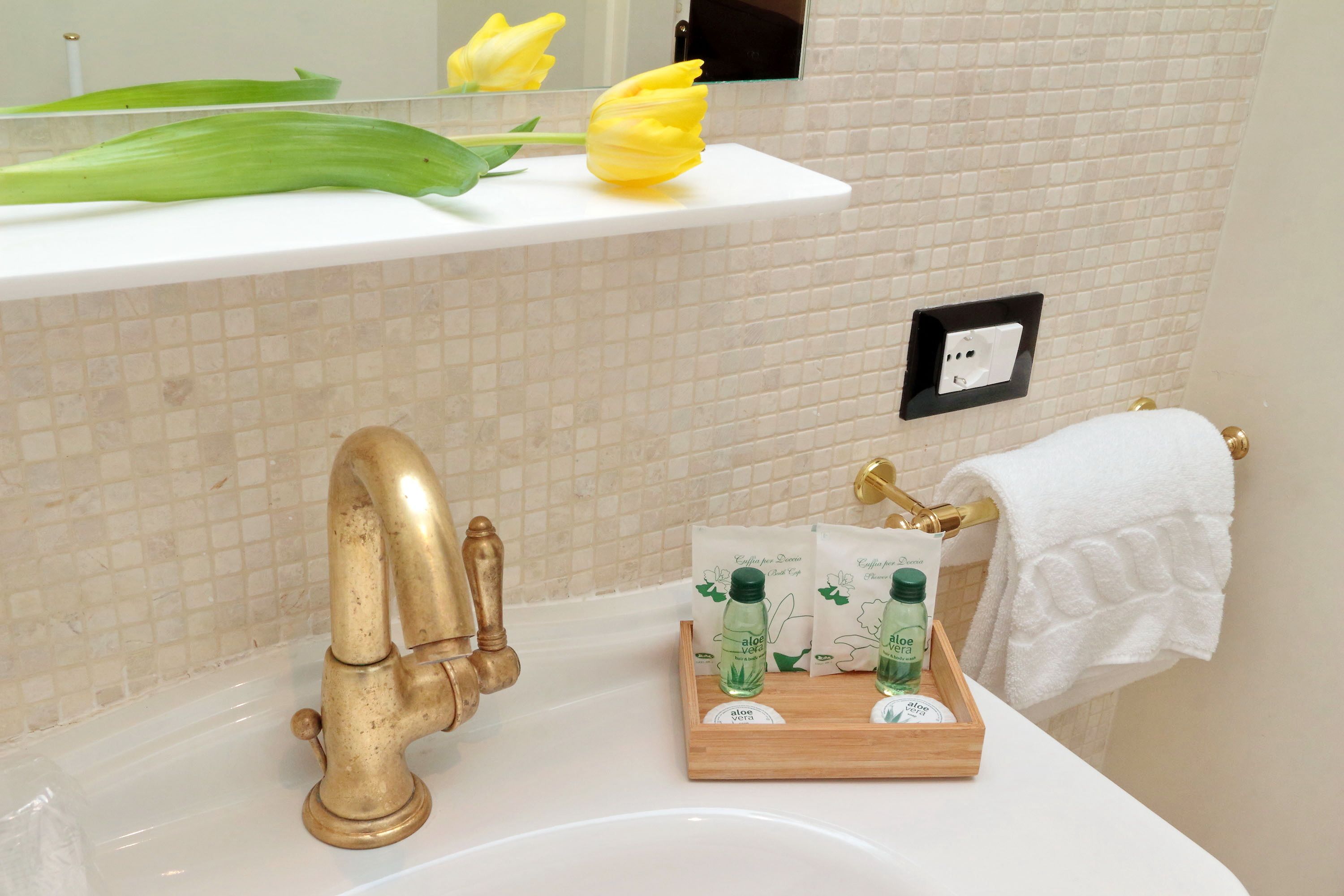Un bagno con un lavandino bianco, un rubinetto dorato, un vassoio di legno con flaconi di aloe vera e un tulipano giallo su uno scaffale bianco.