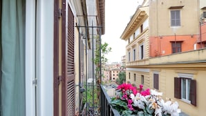 A balcony with a view of adjacent buildings, a metal railing, and potted plants with flowers. - CancelleriaDieci (Rome)