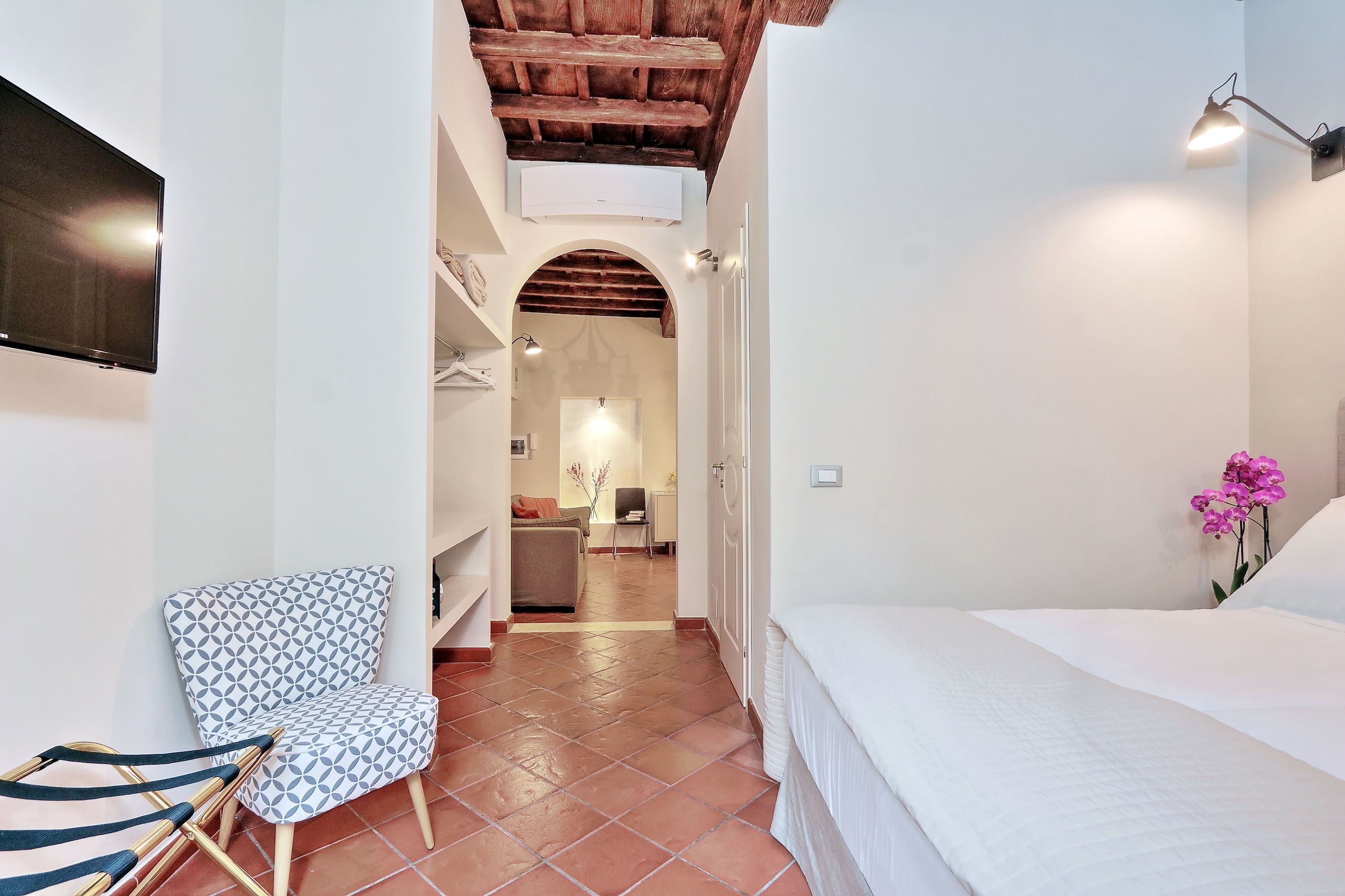 A bedroom with a bed, a chair, a TV, and a hallway leading to a living area.