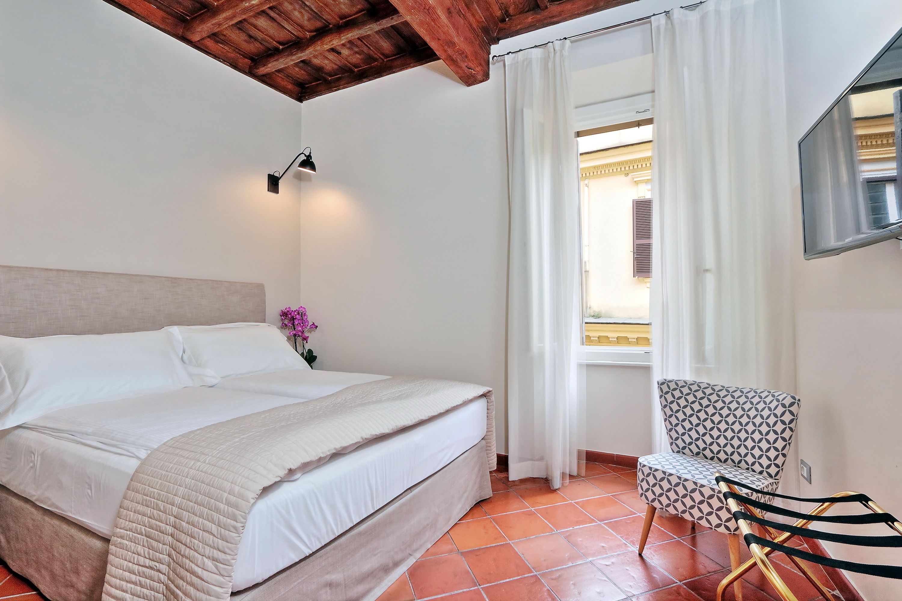 Un dormitorio con una cama, una silla, una ventana con cortinas y un televisor fijado a la pared.