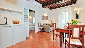 A kitchen with white cabinets, a dining table with chairs, and a view of a living room with a sofa and a painting on the wall. - CancelleriaDieci (Rome)