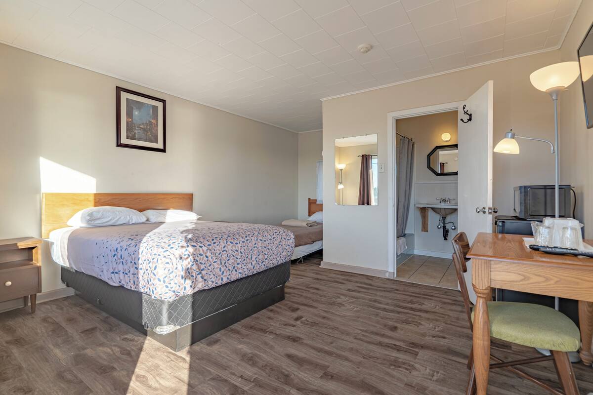 Habitación de hotel con cama, escritorio, silla, espejo y baño visible a través de una puerta abierta.